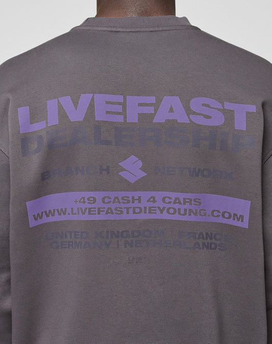 A person wearing the LFDY Network Sweater by LIVE FAST DIE YOUNG—an oversize dark gray sweatshirt with bold purple "LIVEFAST DEALERSHIP" back print, plus details like a website, phone number, and various country names.