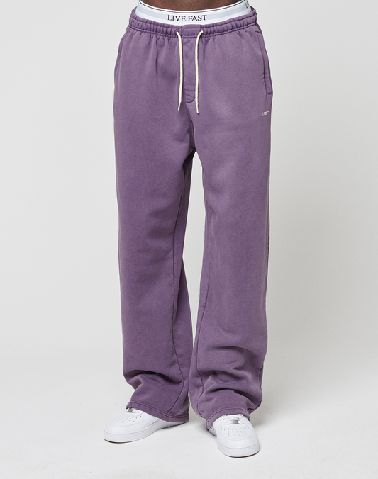 A person wearing the LIVE FAST DIE YOUNG Basic Wide Jogger in purple with a wide fit, elastic waistband, white drawstrings, white sneakers, and a visible LIVE FAST logo-print waistband stands against a plain white background.