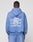 A person wearing the LIVE FAST DIE YOUNG Basic Evidence Hooded, an oversized blue hoodie with a faded finish and repeated LiveFast prints in white and navy, plus overlapping receipts and symbols on the back, faces away from the camera.