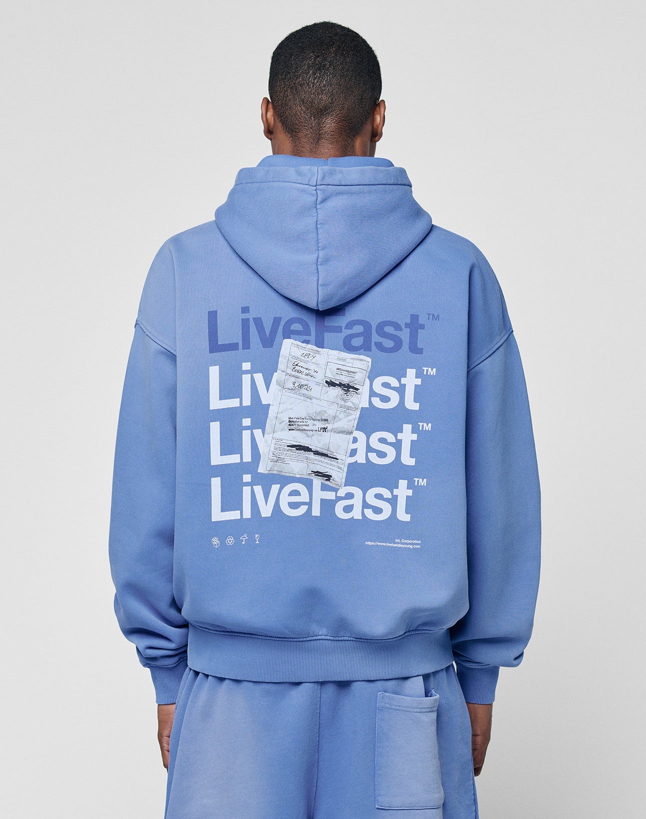 A person wearing the LIVE FAST DIE YOUNG Basic Evidence Hooded, an oversized blue hoodie with a faded finish and repeated LiveFast prints in white and navy, plus overlapping receipts and symbols on the back, faces away from the camera. | model