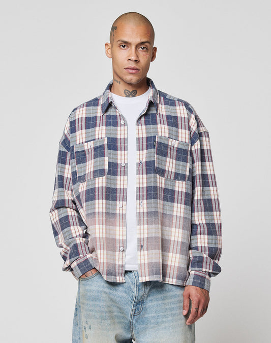 A person with a shaved head wears an oversize fit LIVE FAST DIE YOUNG Vintage Flannel in blue and beige plaid over a white T-shirt and light blue jeans, standing with one hand in their pocket against a plain light gray background.