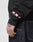 Wearing an oversize fit black LFDY Team Hooded Zip by LIVE FAST DIE YOUNG with UK, Dutch, and German flag patches on the sleeve and blue jeans. Their tattooed hand rests casually by their side.
