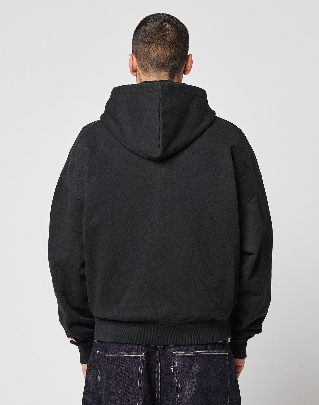 A person with short hair stands with their back to the camera, wearing the LIVE FAST DIE YOUNG LFDY Team Hooded Zip in black with Multilogo Stickereien and dark jeans, set against a plain light grey background.