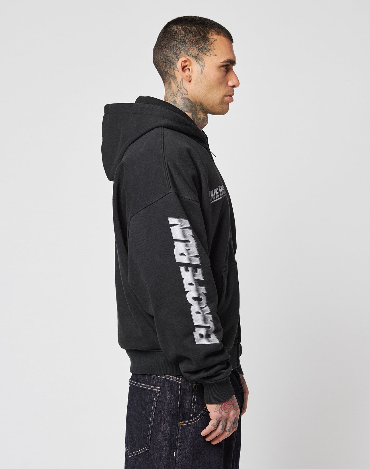 A tattooed person in profile wears the LIVE FAST DIE YOUNG LFDY Team Hooded Zip, an oversize-fit black hoodie with flag embroidery on the sleeve, shown against a plain white background.