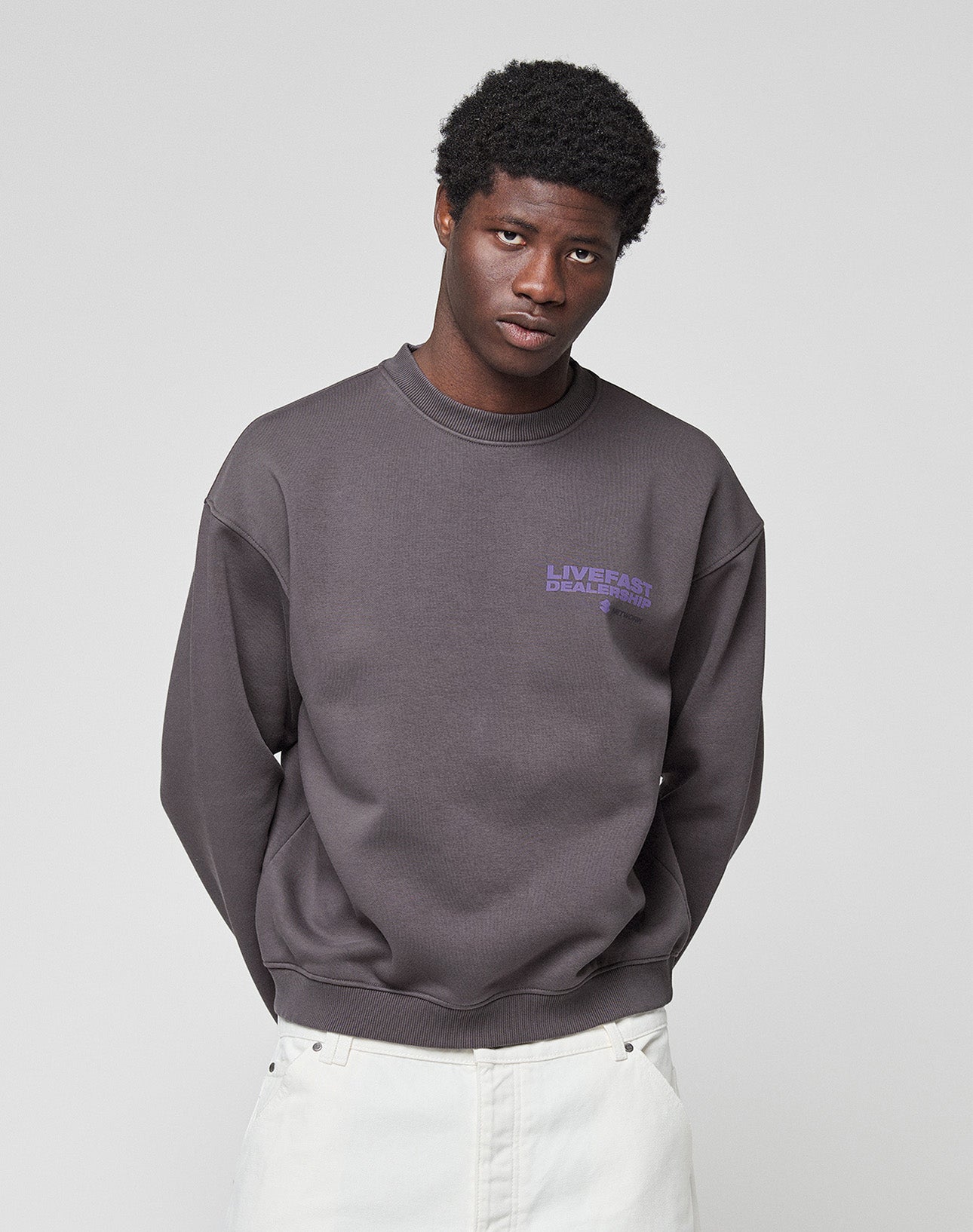 A person wears the oversized LFDY Network Sweater by LIVE FAST DIE YOUNG in dark grey Baumwolle, featuring purple "LIVE FAST DEATH LAST" prints on front and back, standing against a plain light background and facing the camera.