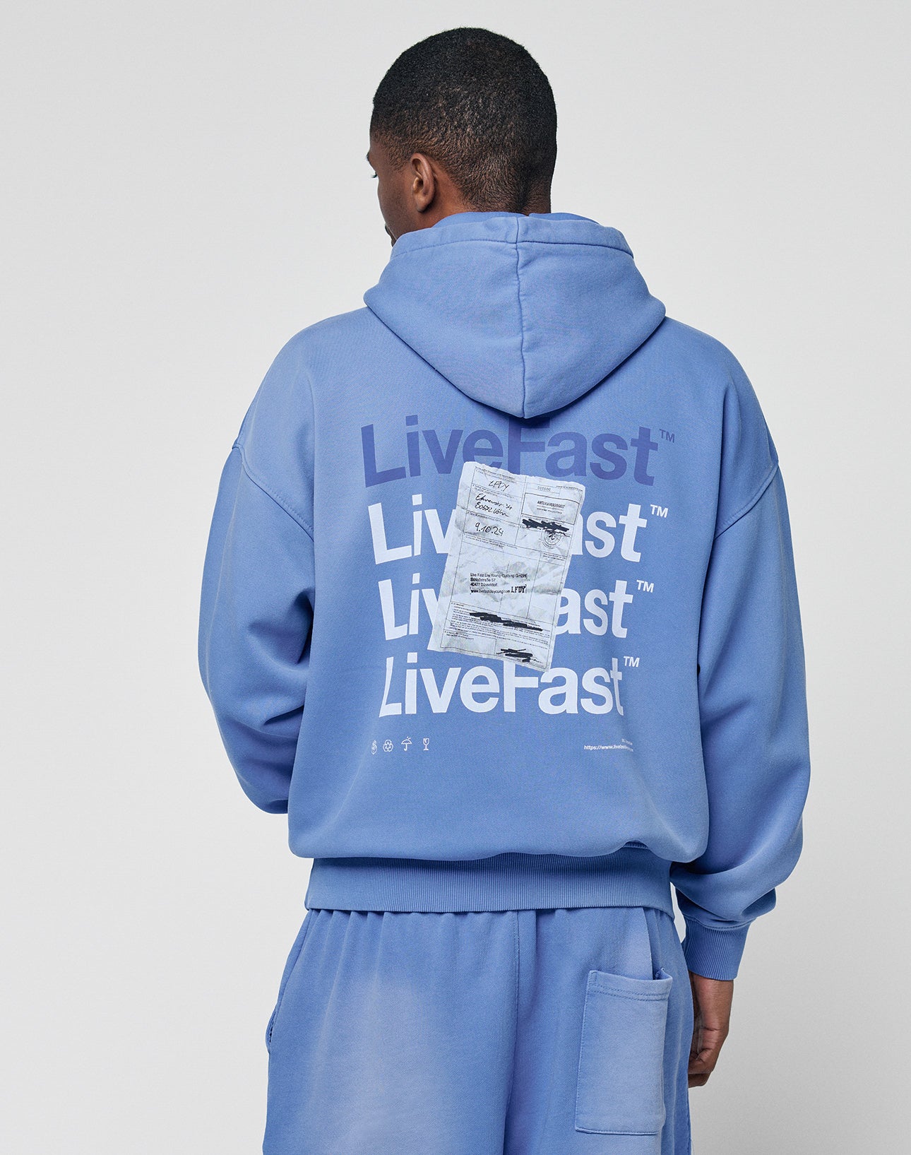 Someone wearing an oversize fit blue Basic Evidence Hooded by LIVE FAST DIE YOUNG with matching shorts stands facing away. The hoodie’s back shows a faded Live Fast print repeated in white, partly covered by a white rectangle graphic.