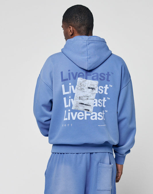 Someone wearing an oversize fit blue Basic Evidence Hooded by LIVE FAST DIE YOUNG with matching shorts stands facing away. The hoodie’s back shows a faded Live Fast print repeated in white, partly covered by a white rectangle graphic.
