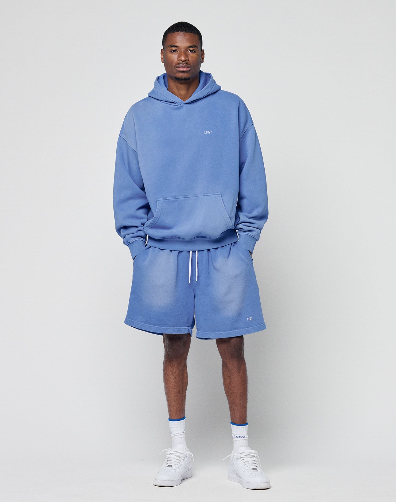 A man stands against a plain background wearing the LIVE FAST DIE YOUNG Basic Evidence Hooded light blue oversized hoodie and shorts set with a subtle faded look, paired with white crew socks and sneakers, looking at the camera with hands in his pockets.