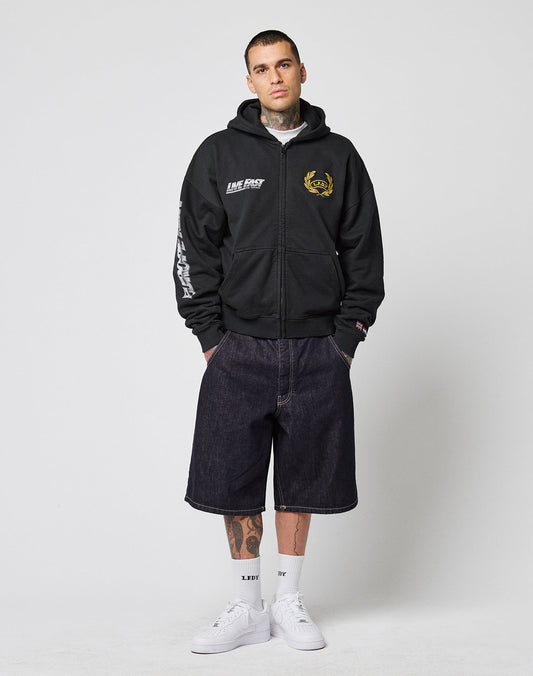 A man stands against a plain backdrop wearing the LIVE FAST DIE YOUNG LFDY Team Hooded Zip (black, oversize fit, multilogo stickereien), dark baggy knee-length shorts, white LIFEDIV socks, white sneakers, and Flaggen-Stick am Ärmel. Hands in pockets.