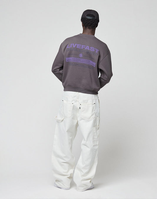 A person stands facing away in a dark cap, white loose pants, and the LIVE FAST DIE YOUNG "LFDY Network Sweater," an oversized purple cotton sweatshirt with LIVEFAST front and back print, against a plain light studio background.