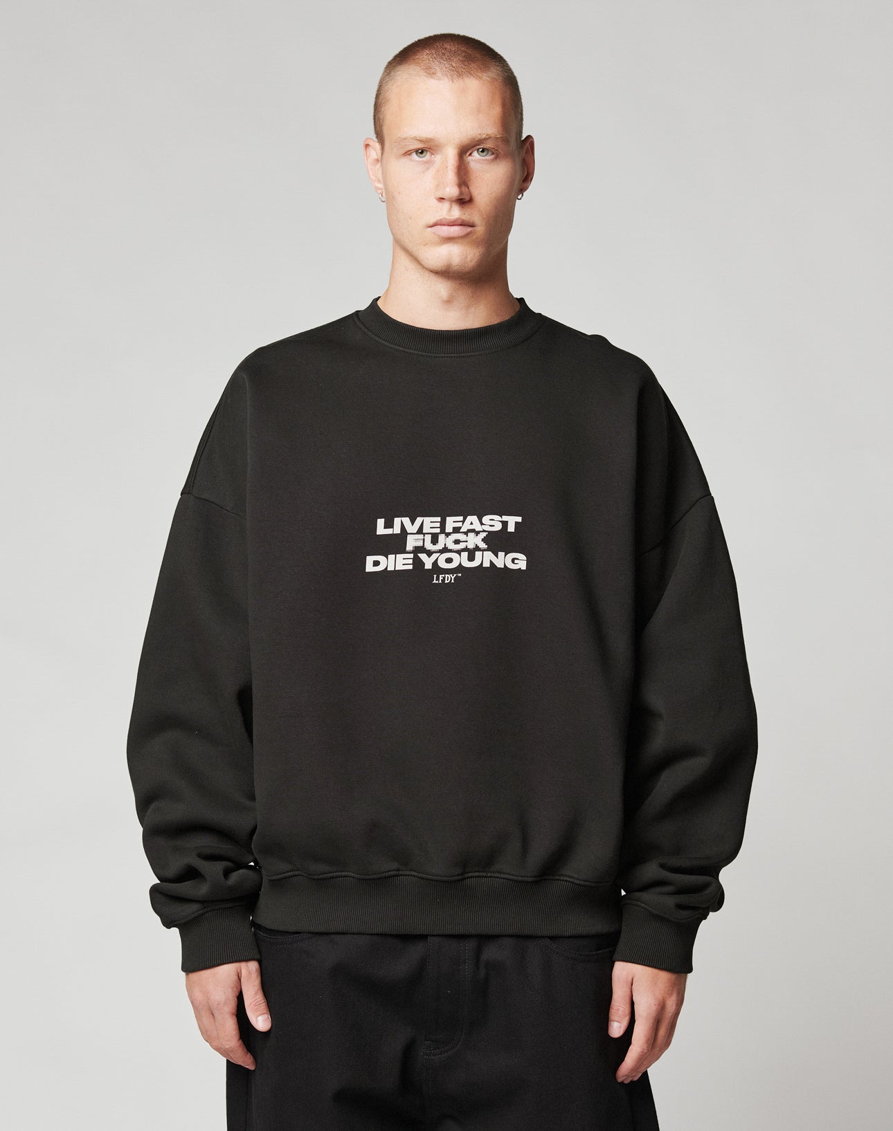 A person with short hair wears the Fuck Die Young Sweater by LIVE FAST DIE YOUNG, an oversize black Baumwolle sweatshirt. Facing forward, they have a neutral expression against a plain light gray background.