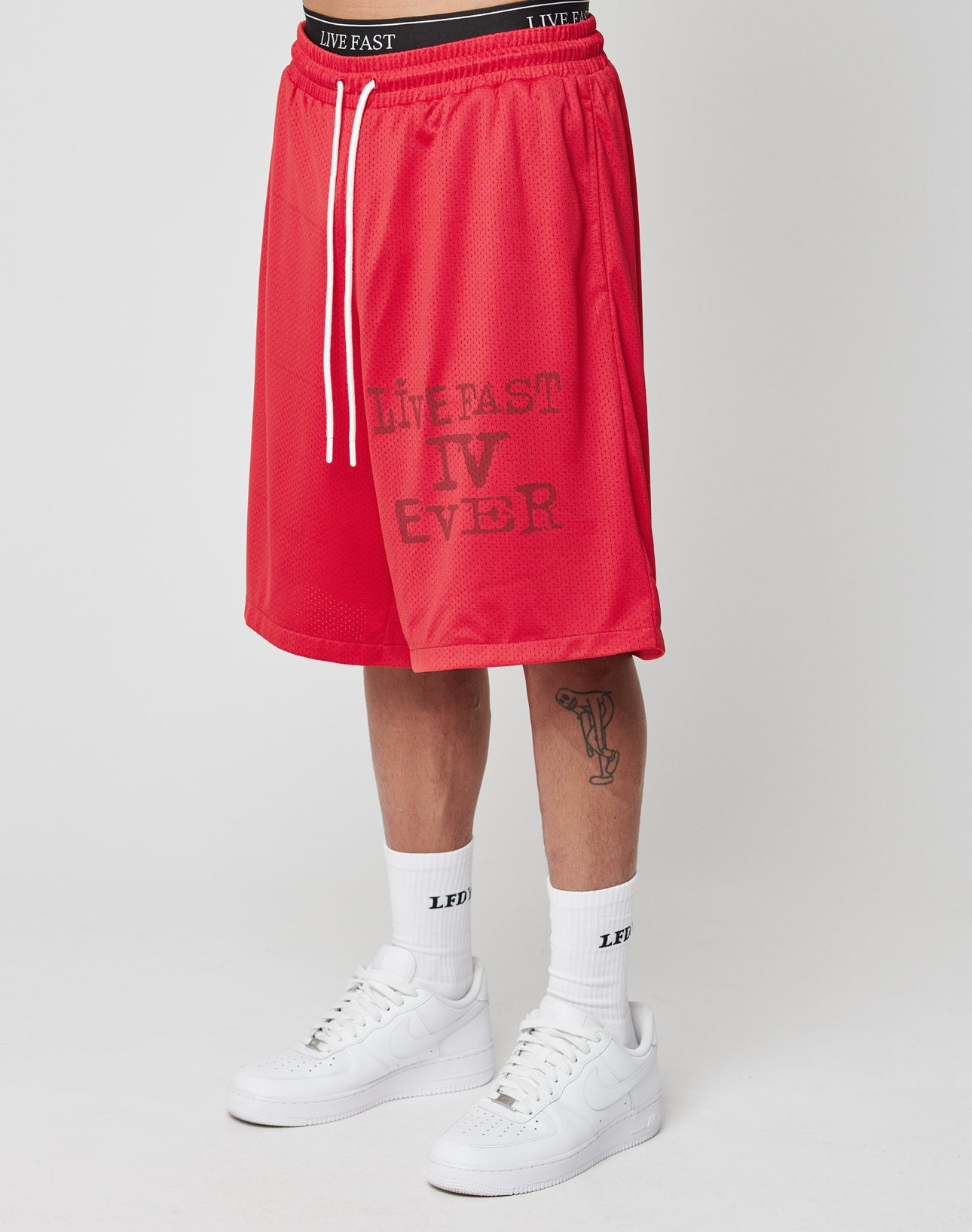 A person wears LIVE FAST DIE YOUNG Live Fast IV Ever Shorts with an elastic waistband and side pockets, featuring LIVE FAST IV EVER print, white LFDY socks, white Nike Air Force 1s, and has a trumpet tattoo on their left shin.