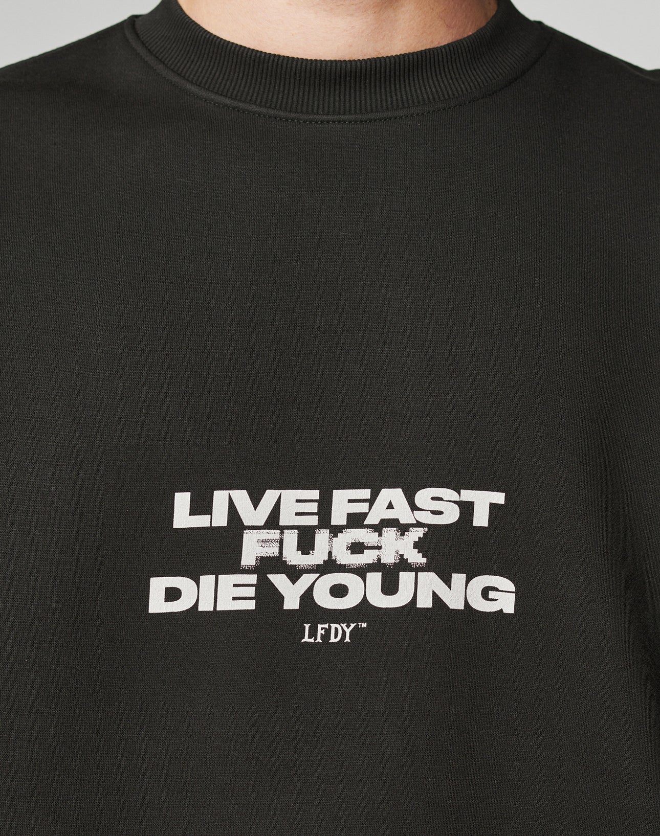 A close-up of the Fuck Die Young Sweater by LIVE FAST DIE YOUNG, an oversized black sweatshirt in soft cotton and polyester, featuring bold white LIVE FAST FUCK DIE YOUNG LFDFY text with a slightly pixelated FUCK on the front.