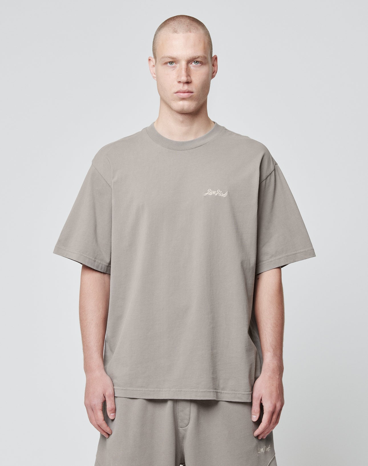 A person with a buzz cut wears the taupe Basic Script Tee by LIVE FAST DIE YOUNG in an oversized fit with matching shorts, standing against a plain light gray background and facing forward with a neutral expression.