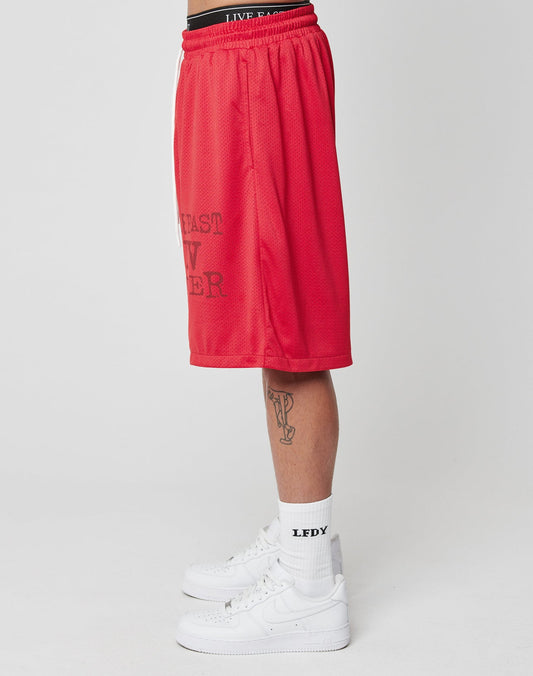 A person in LIVE FAST DIE YOUNG Live Fast IV Ever Shorts, with elastic waistband and side pockets, white LEDY socks, white sneakers, and a visible leg tattoo, stands sideways against a plain white background.
