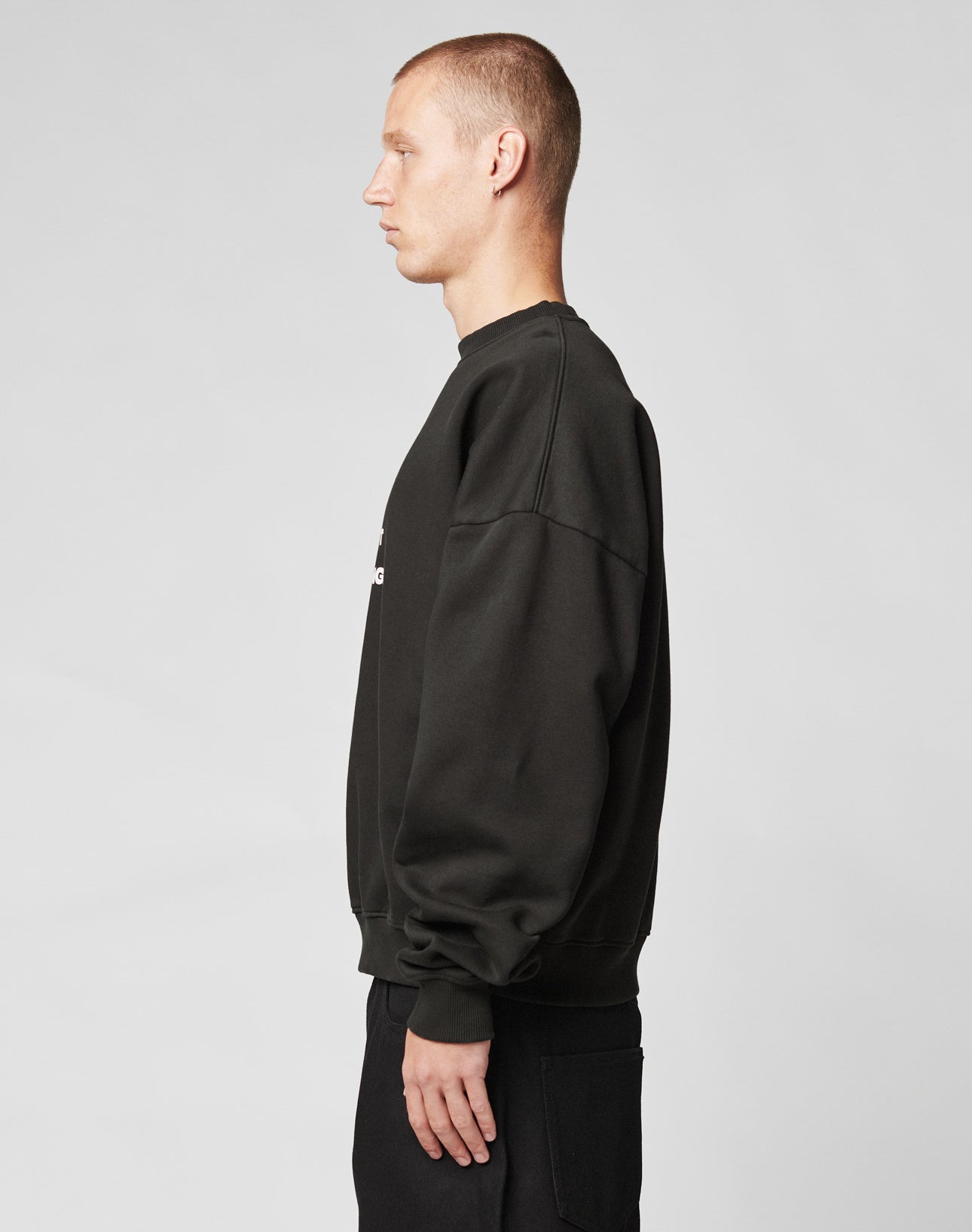 A young man with short hair stands in profile facing left, wearing the LIVE FAST DIE YOUNG Fuck Die Young Sweater in an oversize fit, paired with matching black pants, set against a plain light gray background.