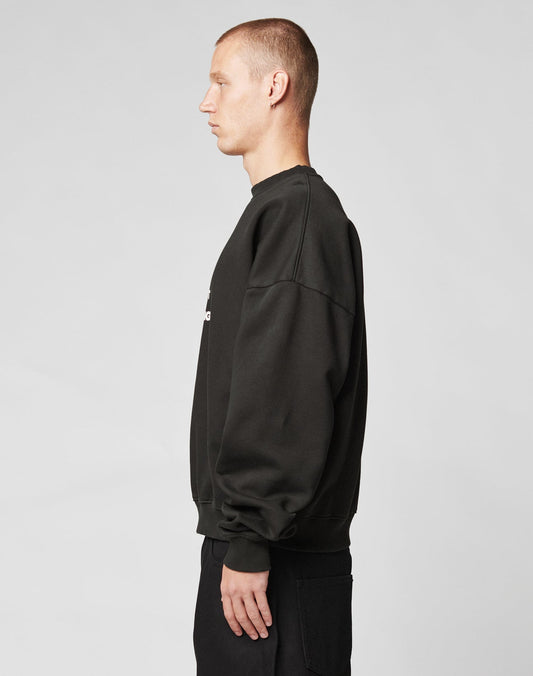A young man with short hair stands in profile facing left, wearing the LIVE FAST DIE YOUNG Fuck Die Young Sweater in an oversize fit, paired with matching black pants, set against a plain light gray background.