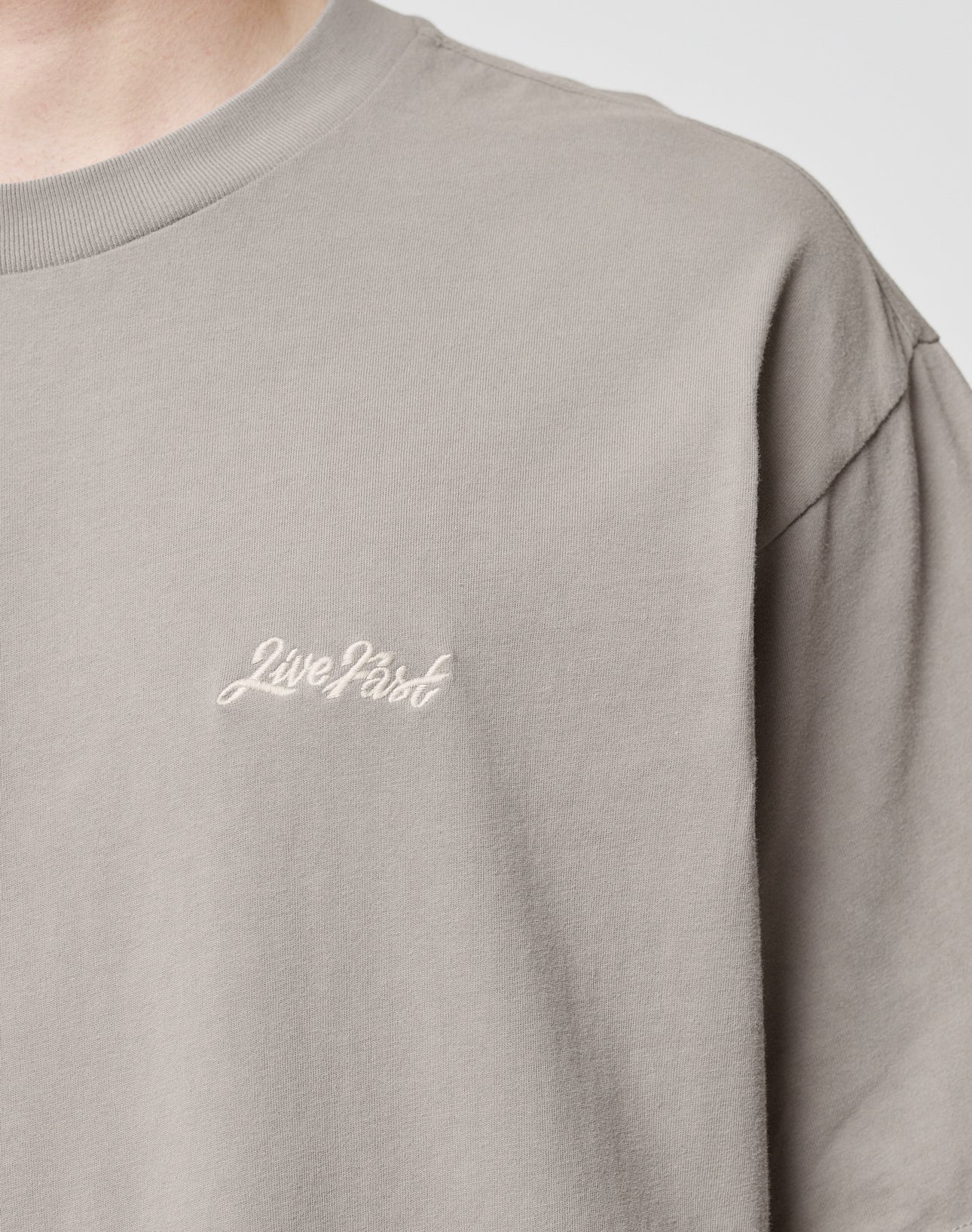 A close-up shows the upper torso and neck of a person wearing the LIVE FAST DIE YOUNG Basic Script Tee in light grey, featuring Live Fast in white script on the chest. Made from soft cotton.