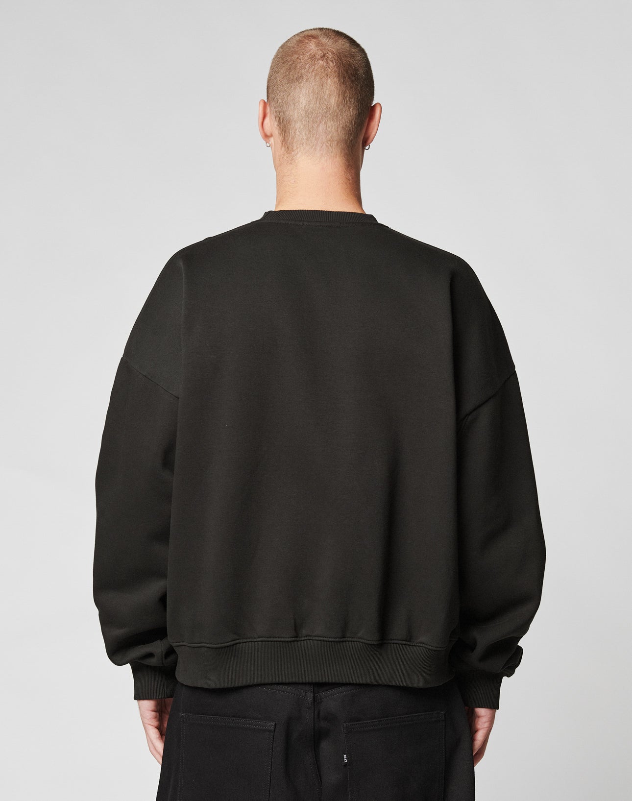 A person with a shaved head is seen from behind wearing the LIVE FAST DIE YOUNG Fuck Die Young Sweater in black, featuring an oversize fit, paired with black pants against a plain light gray background.