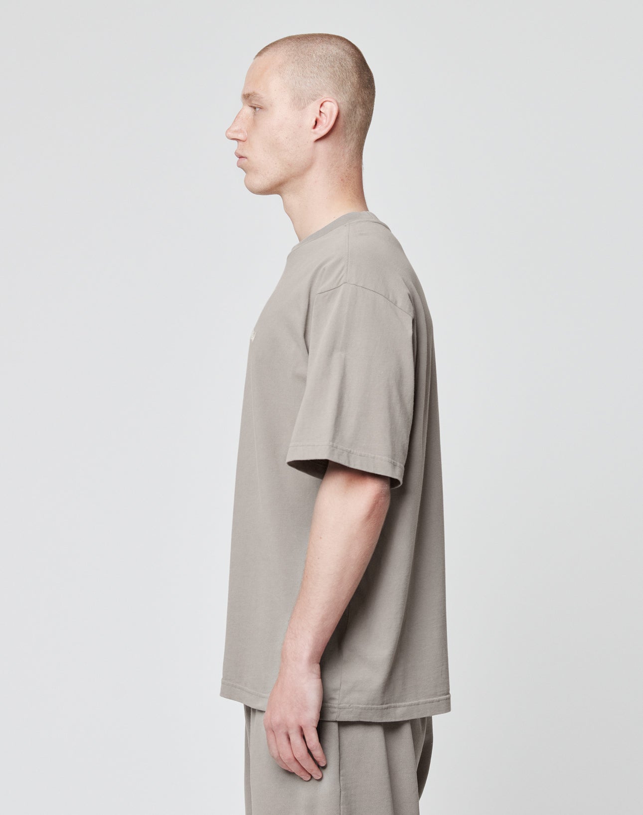 A person with closely shaved hair stands in profile against a plain background, wearing a loose-fitting, short-sleeved taupe Basic Script Tee by LIVE FAST DIE YOUNG with matching pants.