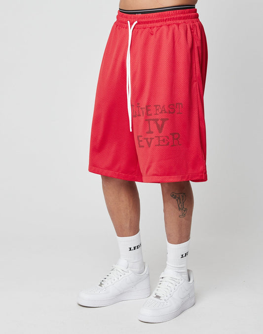 A person wears LIVE FAST DIE YOUNG’s Live Fast IV Ever Shorts with side pockets, branding on the left leg, an elastic waistband, white LEG socks, white sneakers, and a goblet tattoo on their left leg against a plain background.