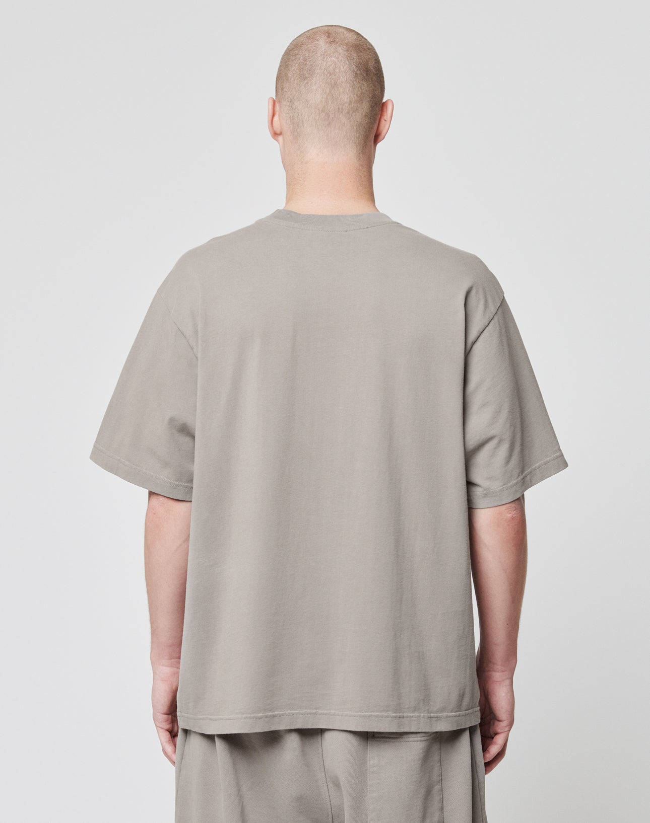 A person with a shaved head stands with their back to the camera, wearing the LIVE FAST DIE YOUNG Basic Script Tee in taupe—loose-fitting, short-sleeve cotton with vintage wash—and matching pants, set against a plain light gray background.