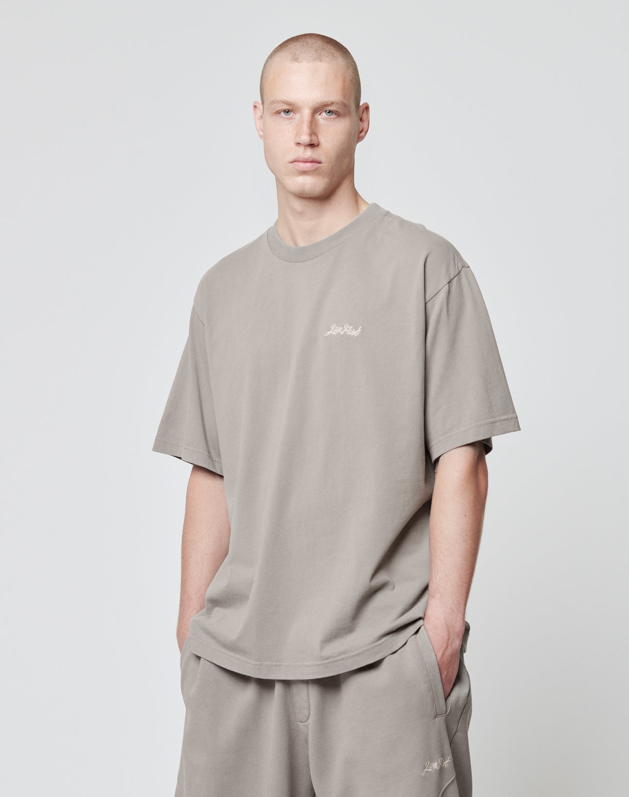 A person with short hair wears the LIVE FAST DIE YOUNG Basic Script Tee and matching shorts in an Oversize Fit, light gray color, standing against a plain background with hands in their pockets.