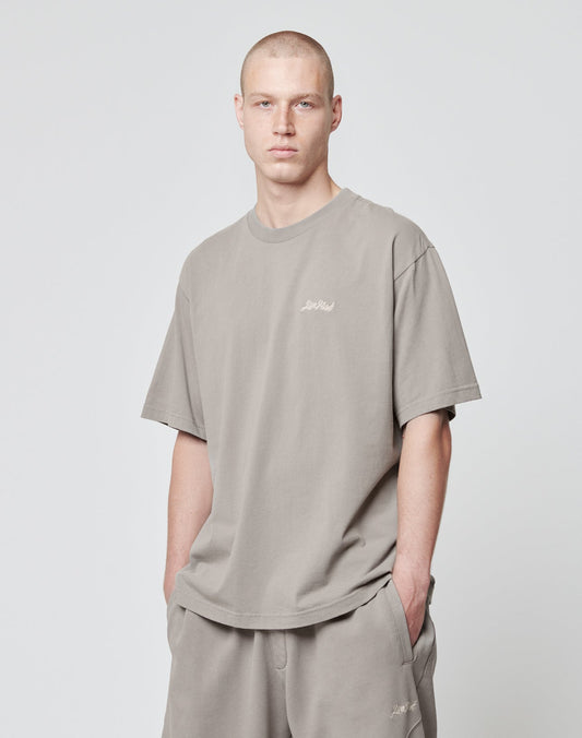 A person with short hair wears the LIVE FAST DIE YOUNG Basic Script Tee and matching shorts in an Oversize Fit, light gray color, standing against a plain background with hands in their pockets.