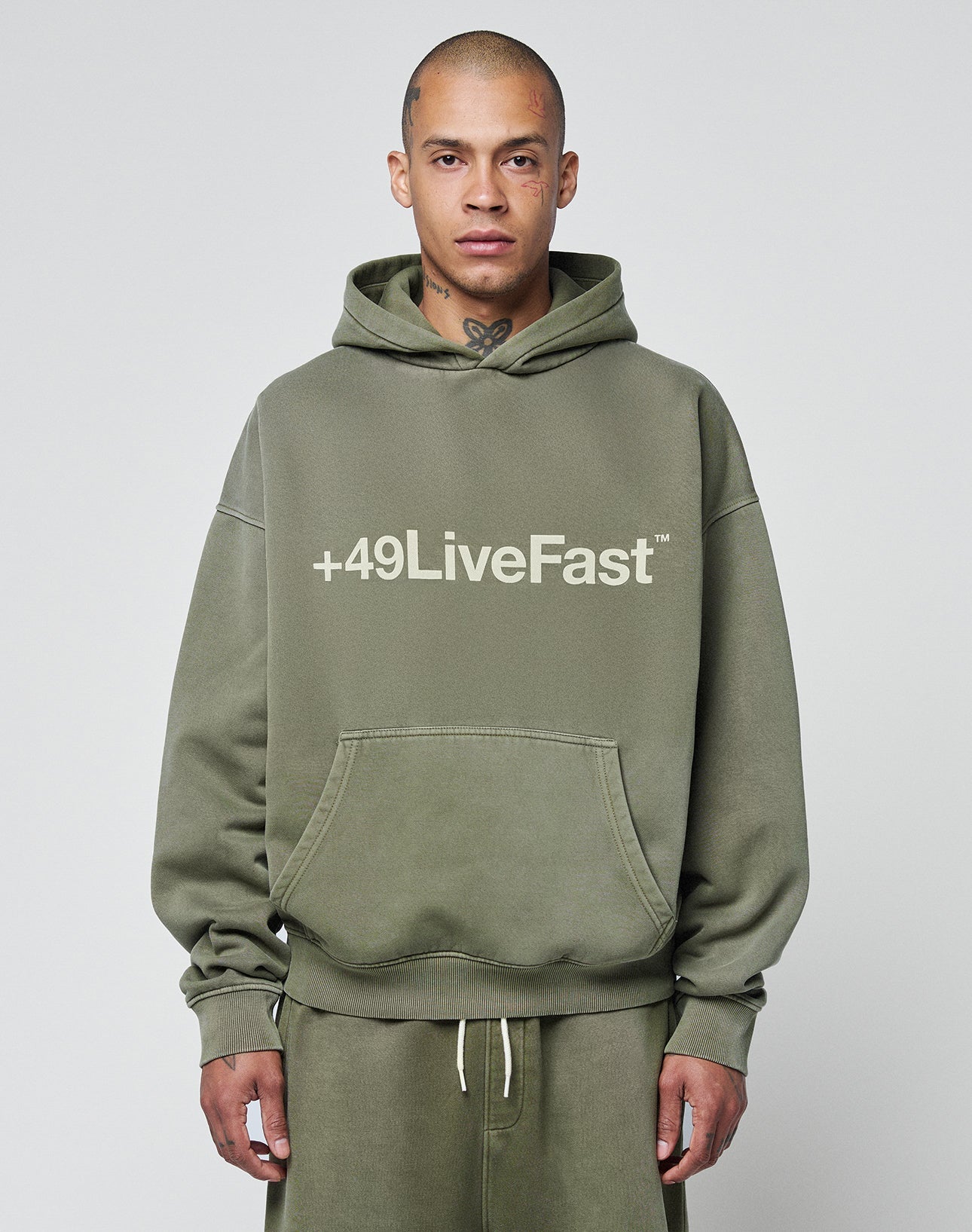 A person with a shaved head and neck tattoo wears the Basic +49 Hooded by LIVE FAST DIE YOUNG—an olive green, oversized hoodie with +49LiveFast on the front, paired with matching faded sweatpants, standing against a plain light gray background.
