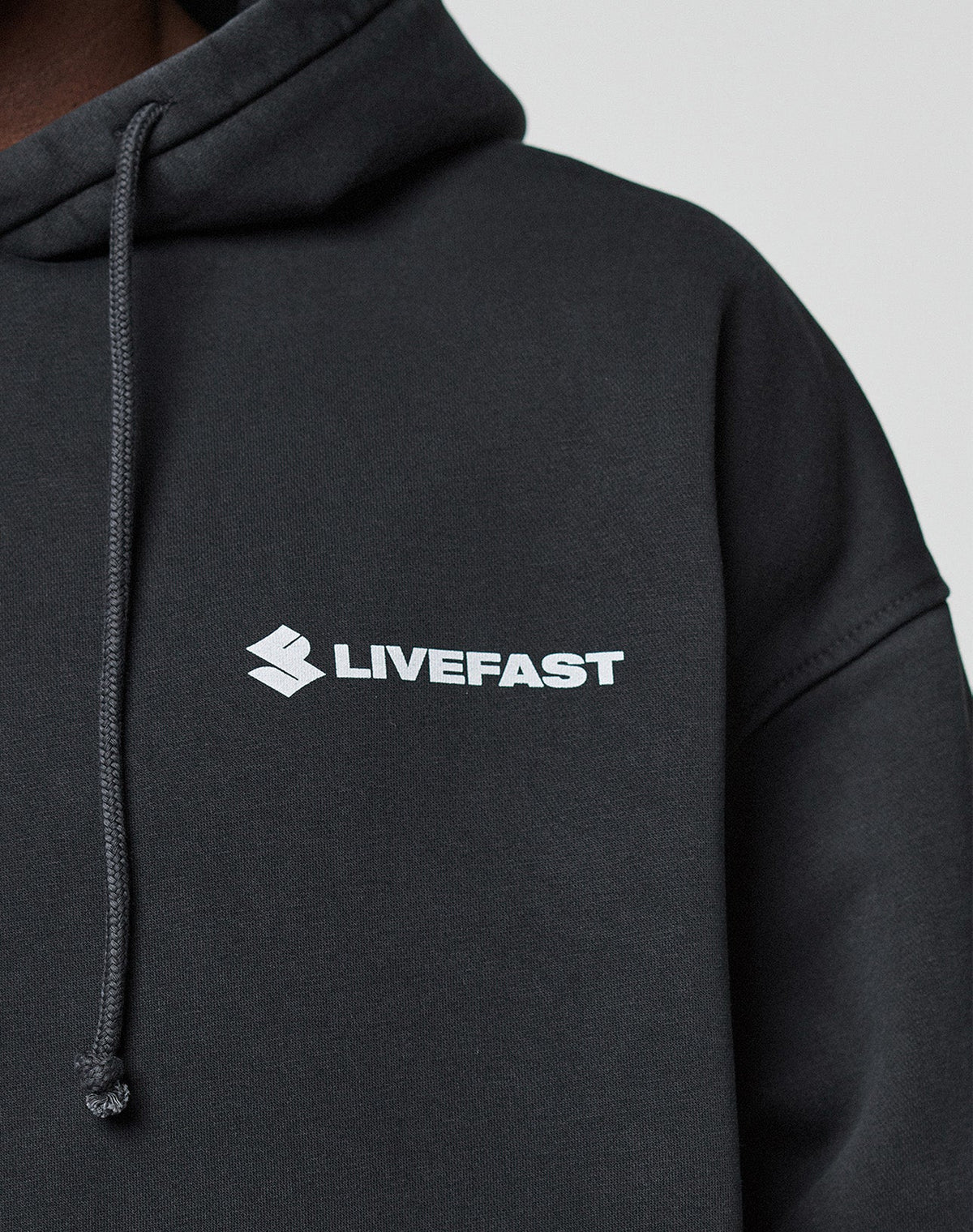 A close-up shows a person in the black LFDY Dealership Hooded by LIVE FAST DIE YOUNG, made from Baumwolle, with a white LIVEFAST logo and emblem on the chest. The oversized hoodie has a drawstring and is set against a light background.