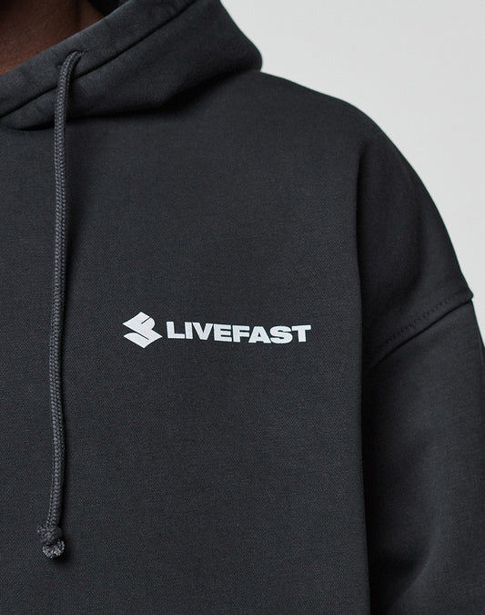 A close-up shows a person in the black LFDY Dealership Hooded by LIVE FAST DIE YOUNG, made from Baumwolle, with a white LIVEFAST logo and emblem on the chest. The oversized hoodie has a drawstring and is set against a light background.