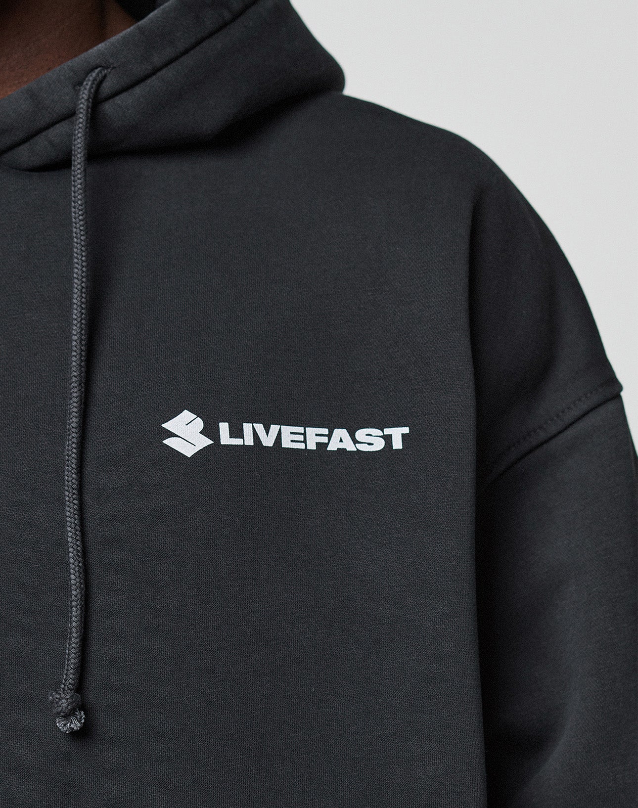 A close-up shows a person in the black LFDY Dealership Hooded by LIVE FAST DIE YOUNG, made from Baumwolle, with a white LIVEFAST logo and emblem on the chest. The oversized hoodie has a drawstring and is set against a light background.