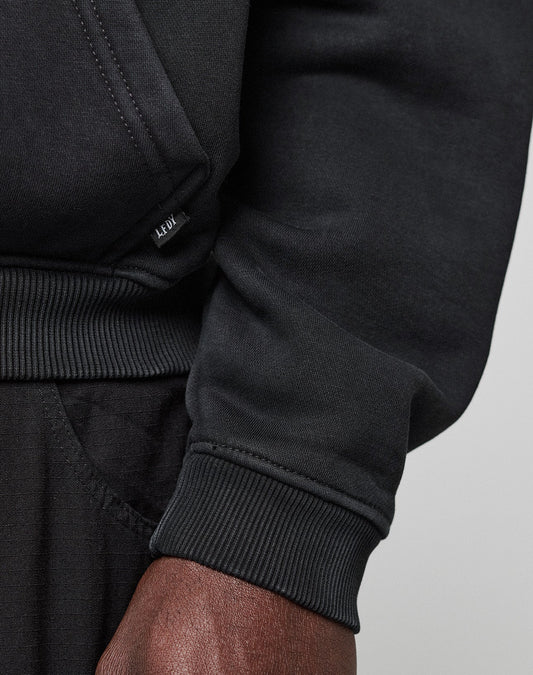 Close-up of a person in a black LFDY Dealership Hooded sweatshirt and black pants. Focus on the ribbed cuff, hand, and small LEDU label. Crafted by LIVE FAST DIE YOUNG from soft cotton for ultimate comfort.
