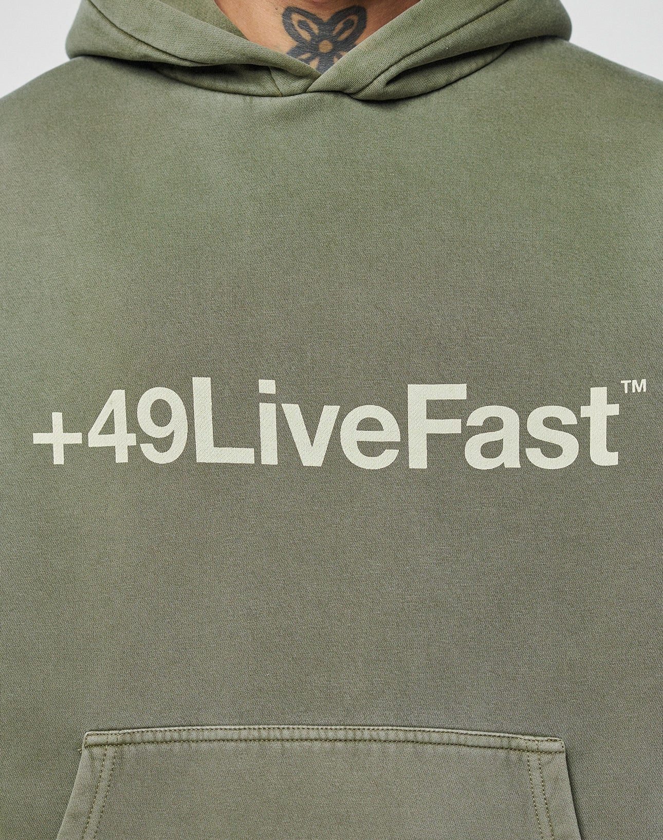 A close-up of the olive-green Basic +49 Hooded by LIVE FAST DIE YOUNG features a bold white +49LiveFast™ print across the chest. The oversize fit adds comfort, with part of a neck tattoo visible at the top of the image.