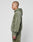 A person with short hair and visible face tattoos stands in profile against a light background, wearing an oversized olive green LIVE FAST DIE YOUNG Basic +49 Hooded hoodie and matching faded pants.