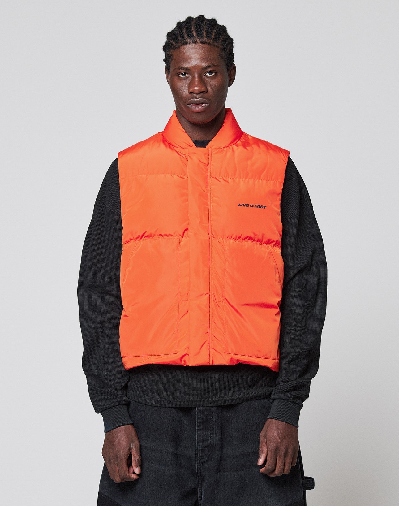 A man wears the LIVE FAST DIE YOUNG Live Fast Cropped Vest in orange with Druckknöpfe over a black sweatshirt and dark pants, standing against a plain light gray background and looking directly at the camera.