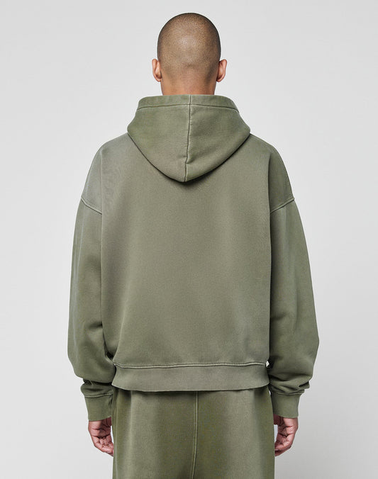 A person with a shaved head models the LIVE FAST DIE YOUNG Basic +49 Hooded sweatshirt and matching pants in olive green, featuring an oversize fit and subtle faded look, standing with their back to the camera against a light gray background.