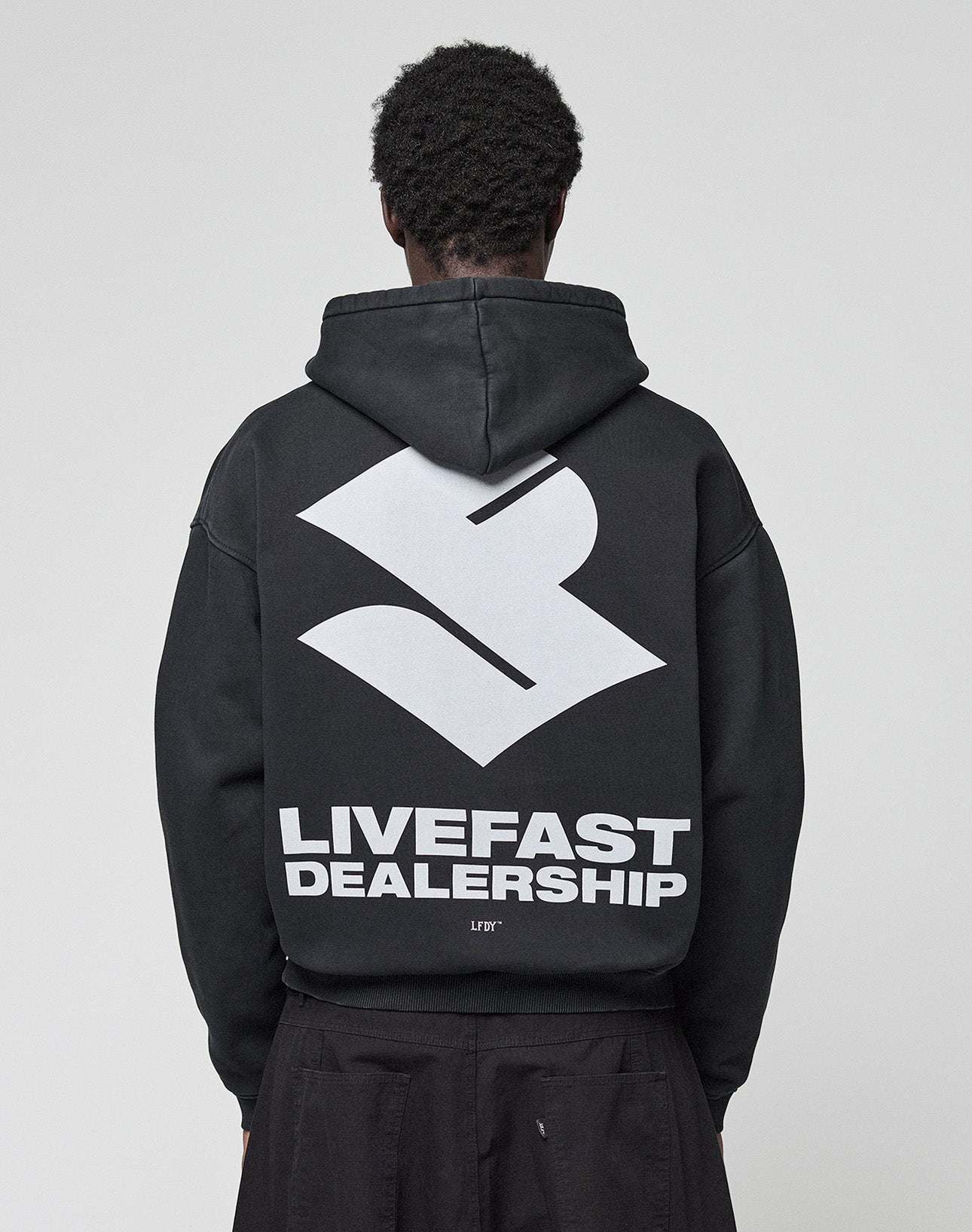 A person with short, curly hair is seen from behind in the black LFDY Dealership Hooded by LIVE FAST DIE YOUNG, featuring an oversize fit with a large white geometric logo and bold LIVEFAST DEALERSHIP text as a backprint. | model