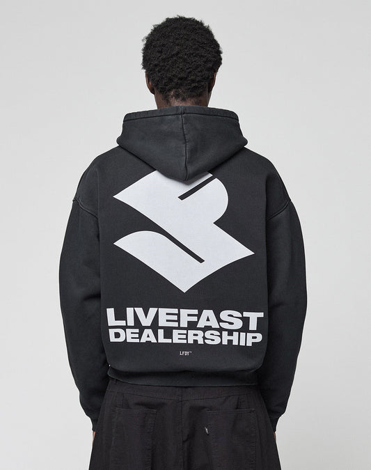 A person with short, curly hair is seen from behind in the black LFDY Dealership Hooded by LIVE FAST DIE YOUNG, featuring an oversize fit with a large white geometric logo and bold LIVEFAST DEALERSHIP text as a backprint. | model