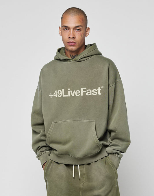 A person in an olive green, oversized LIVE FAST DIE YOUNG Basic +49 Hooded and matching sweatpants with a relaxed, faded look; the hoodie features bold white +49LiveFast text on the front. The person has face and neck tattoos.