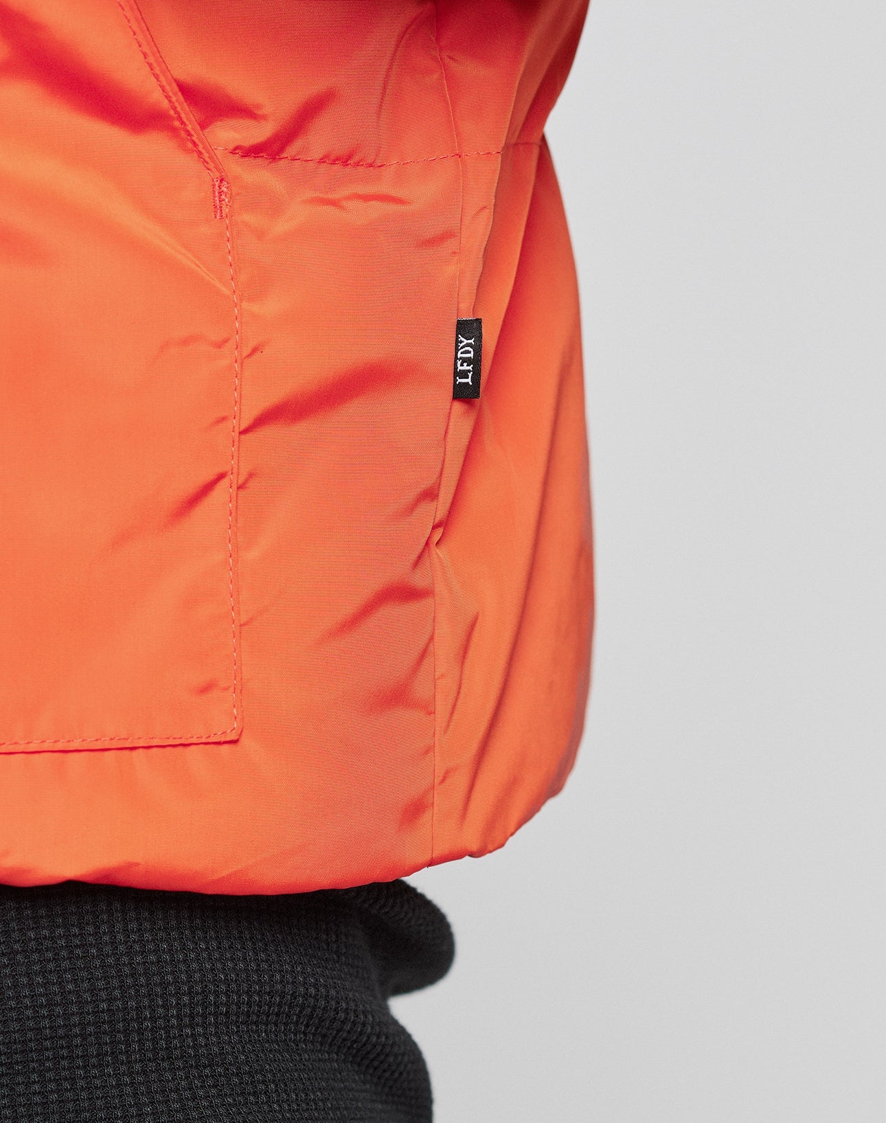 Close-up of the Live Fast Cropped Vest by LIVE FAST DIE YOUNG—an orange jacket with snap buttons and a small black FREY tag near the seam, layered over a black textured top.
