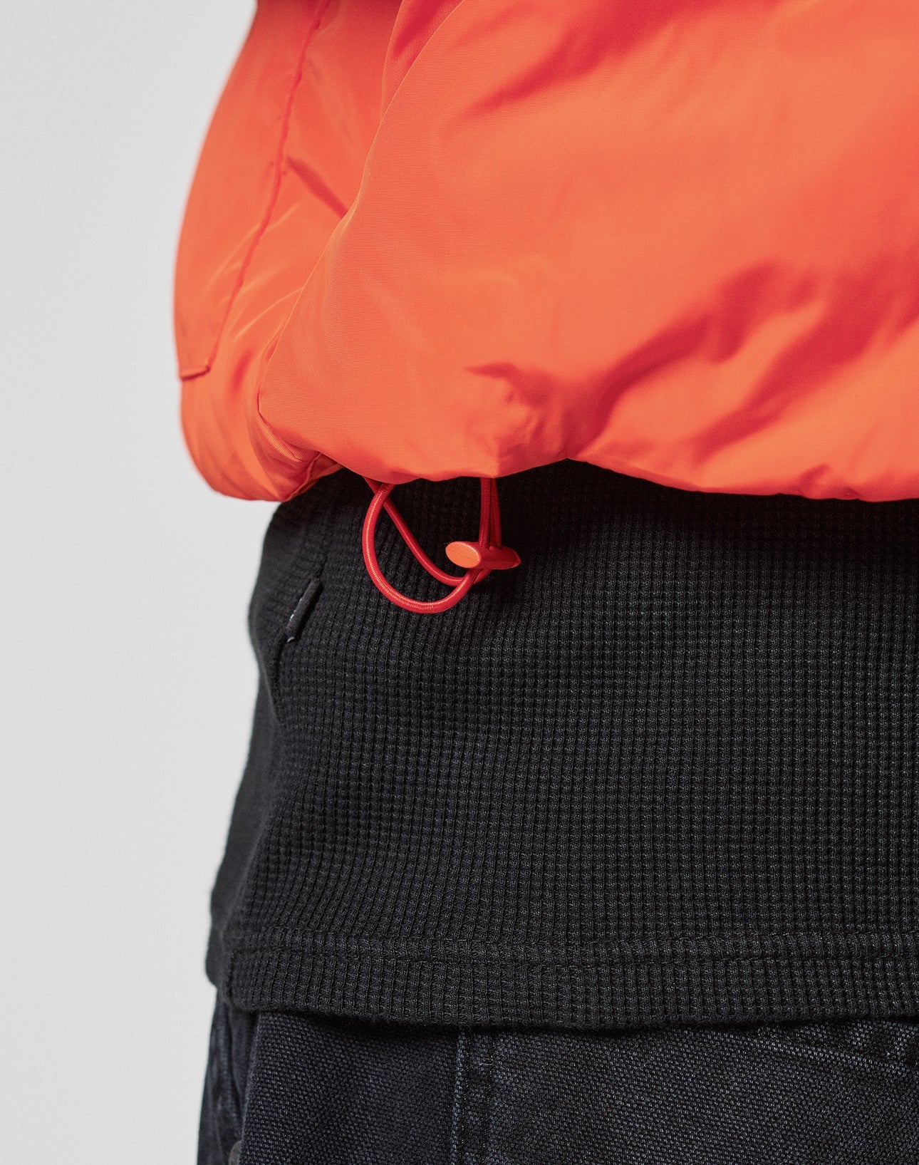 Close-up of a person wearing the Live Fast Cropped Vest by LIVE FAST DIE YOUNG, a bright orange puffer with a red drawstring at the hem, styled over a black ribbed sweater and dark pants. Regular fit with practical Druckknöpfe for easy fastening.