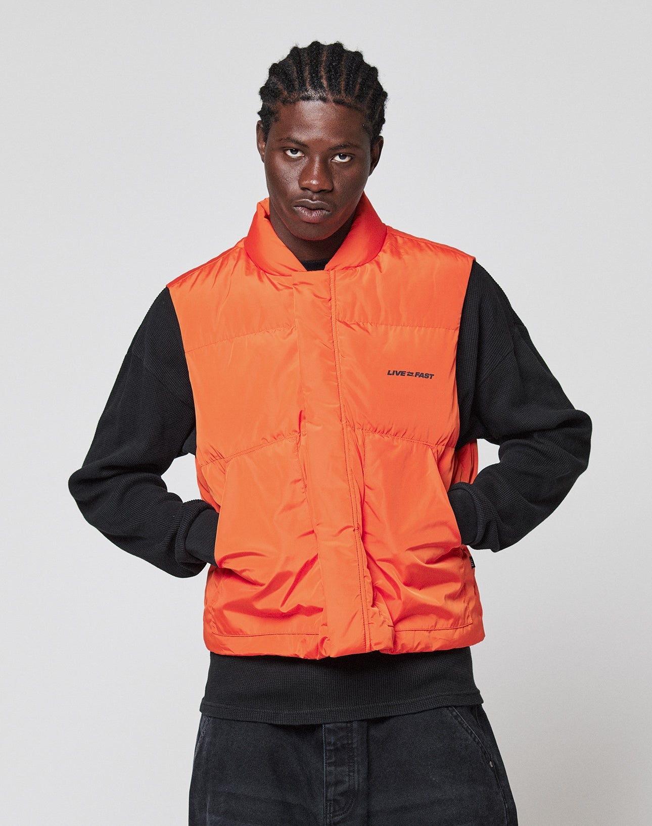 A person with braided hair wears the LIVE FAST DIE YOUNG Live Fast Cropped Vest in bright orange over a black sweatshirt, standing against a plain background with hands in vest pockets. The vest features small “LIFE IS FAST” text on the left side.