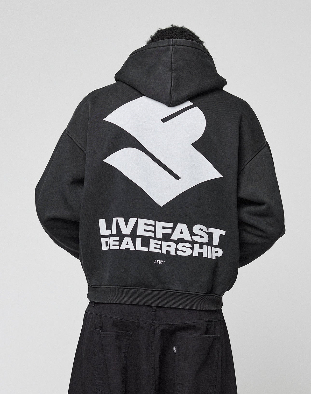 A person wears the LIVE FAST DIE YOUNG LFDY Dealership Hooded, an oversize black Baumwolle hoodie with a bold white abstract logo and "LIVEFAST DEALERSHIP" backprint, photographed from behind against a plain light background.