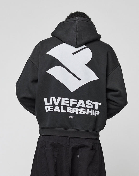 A person wears the LIVE FAST DIE YOUNG LFDY Dealership Hooded, an oversize black Baumwolle hoodie with a bold white abstract logo and "LIVEFAST DEALERSHIP" backprint, photographed from behind against a plain light background.