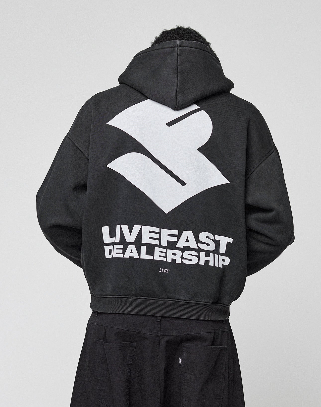 A person wears the LIVE FAST DIE YOUNG LFDY Dealership Hooded, an oversize black Baumwolle hoodie with a bold white abstract logo and "LIVEFAST DEALERSHIP" backprint, photographed from behind against a plain light background.