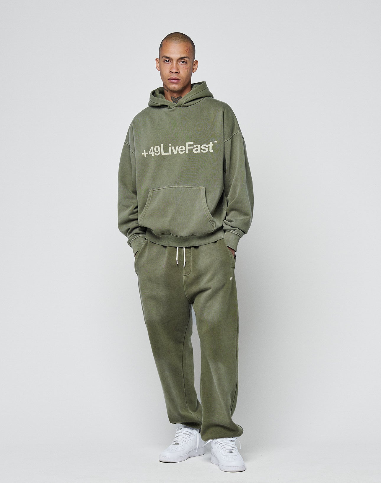 A person stands against a plain light background wearing an olive green LIVE FAST DIE YOUNG hoodie, matching Basic Loose Jogger sweatpants, and white sneakers.