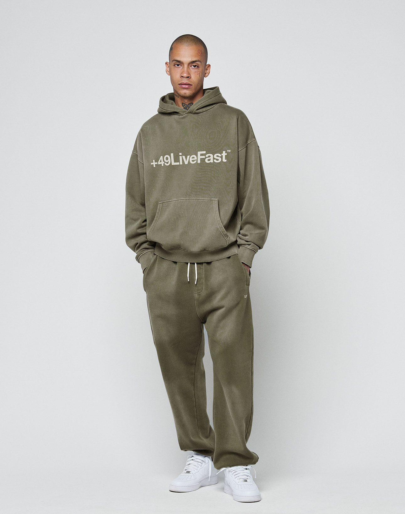 A person with short hair and a neutral expression stands against a plain background, wearing LIVE FAST DIE YOUNG's olive green Basic Loose Jogger and matching hoodie with handgefertigtem Fade-Effekt, paired with white sneakers.