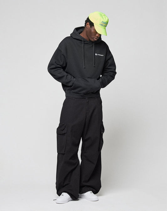 A person in a black LIVE FAST DIE YOUNG LFDY Dealership Hooded with front and back print, black cargo pants, white sneakers, and a neon green cap stands against a light background, hands in the hoodie pocket and gazing slightly down.