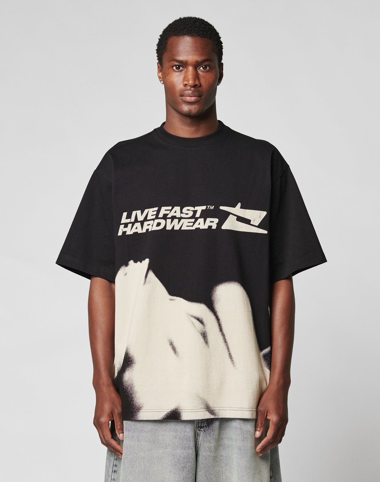 A man faces forward in a black LFDY Mystic Tee by LIVE FAST DIE YOUNG with LIVE FAST HARDWEAR and graphic prints on the front and back, paired with light gray denim shorts. The background is plain and light-colored.